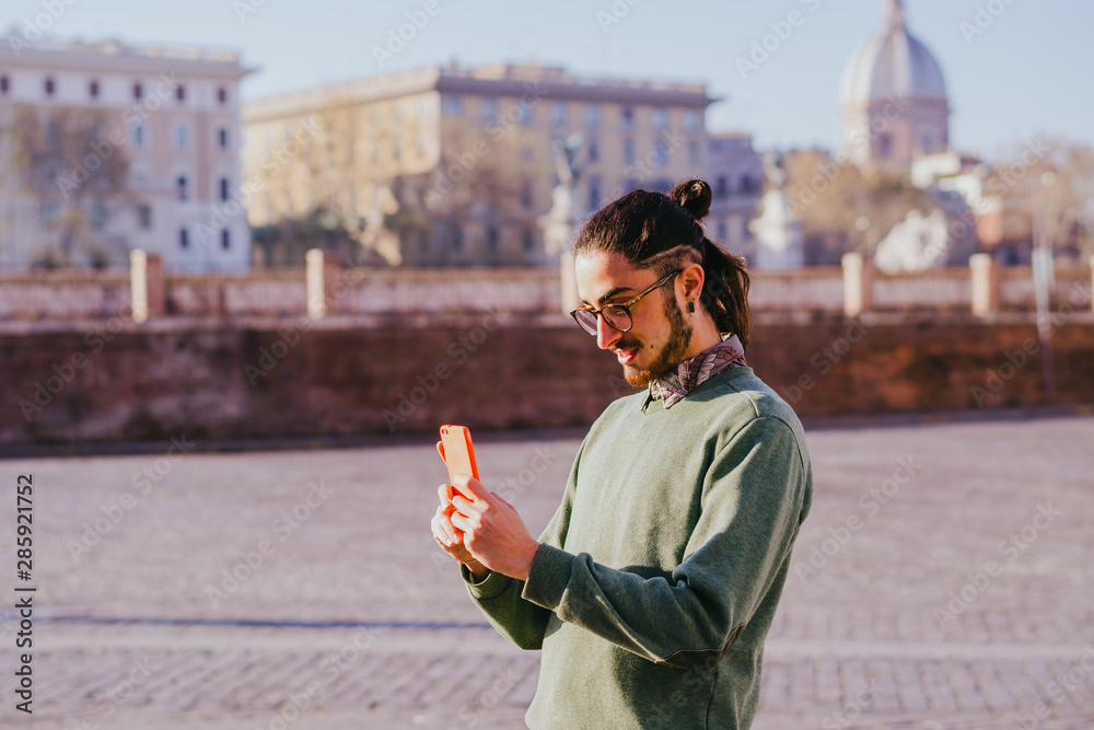 Fototapeta premium Handsome bearded male hipster making photo for blog sharing positive emotions feeling excited by sunny morning while walking to work using modern smartphone and 4G internet