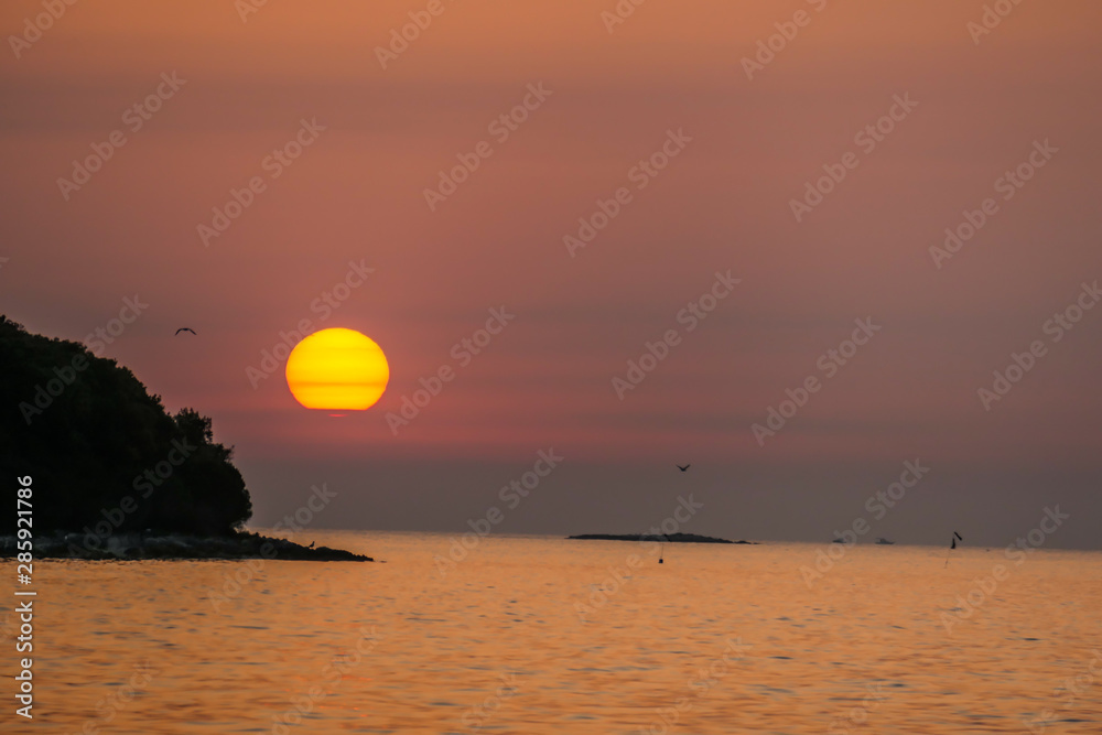 Romantic sunset by a beach. The sun sets over the horizon. The sun beams reflecting in the calm sea waters. There is an island on the side. Few birds flying around. The sky turns yellow and orange Romantic sunset by a beach. The sun sets over the horizon. The sun beams reflecting in the calm sea waters. There is an island on the side. Few birds flying around. The sky turns yellow and orange