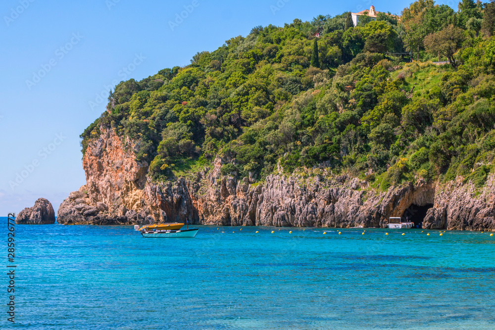Seascape – lagoon with turquoise water, mountain with caves and cliffs, green trees, blooming bushes, rocks in a blue water, colorful cruise touristic boats. Corfu Island, Greece. 
