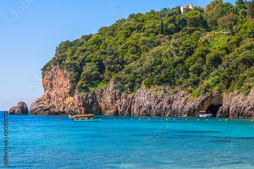 Seascape     lagoon with turquoise water  mountain with caves and cliffs  green trees  blooming bushes  rocks in a blue water  colorful cruise touristic boats. Corfu Island  Greece. 