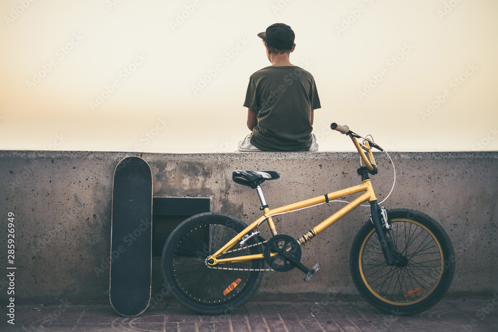 Back view of a teen sitting on a wall with bicycle and skateboard. Young  rider relaxing