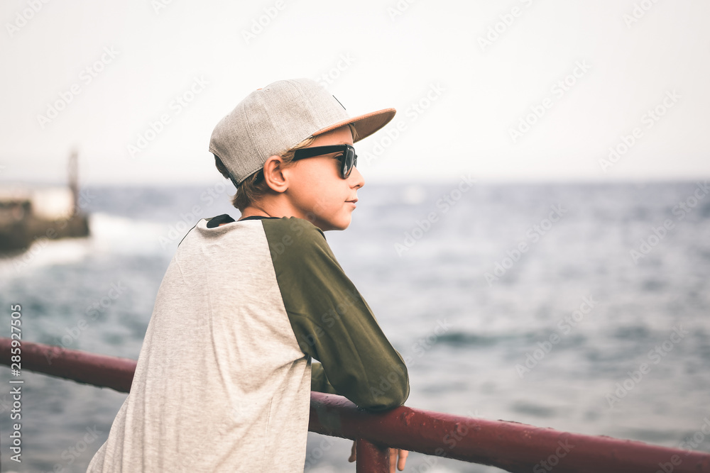 Beautiful smiling teen relaxing leaning against a railing near the sea, looking away with sun glasses and trendy hat. Young boy enjoying last summer days in front of the ocean. Youth, positive concept Beautiful smiling teen relaxing leaning against a railing near the sea, looking away with sun glasses and trendy hat. Young boy enjoying last summer days in front of the ocean. Youth, positive concept