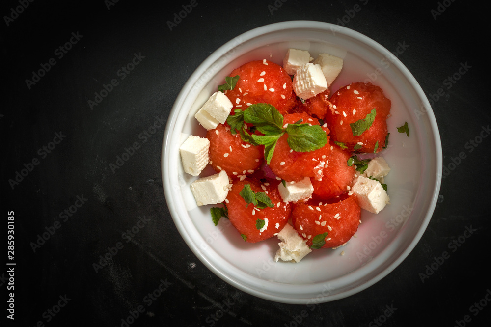 Salad of watermelon balls, feta cheese and mint, sprinkled with sesame seeds in a white plate on a black background. Salad of watermelon balls, feta cheese and mint, sprinkled with sesame seeds in a white plate on a black background.