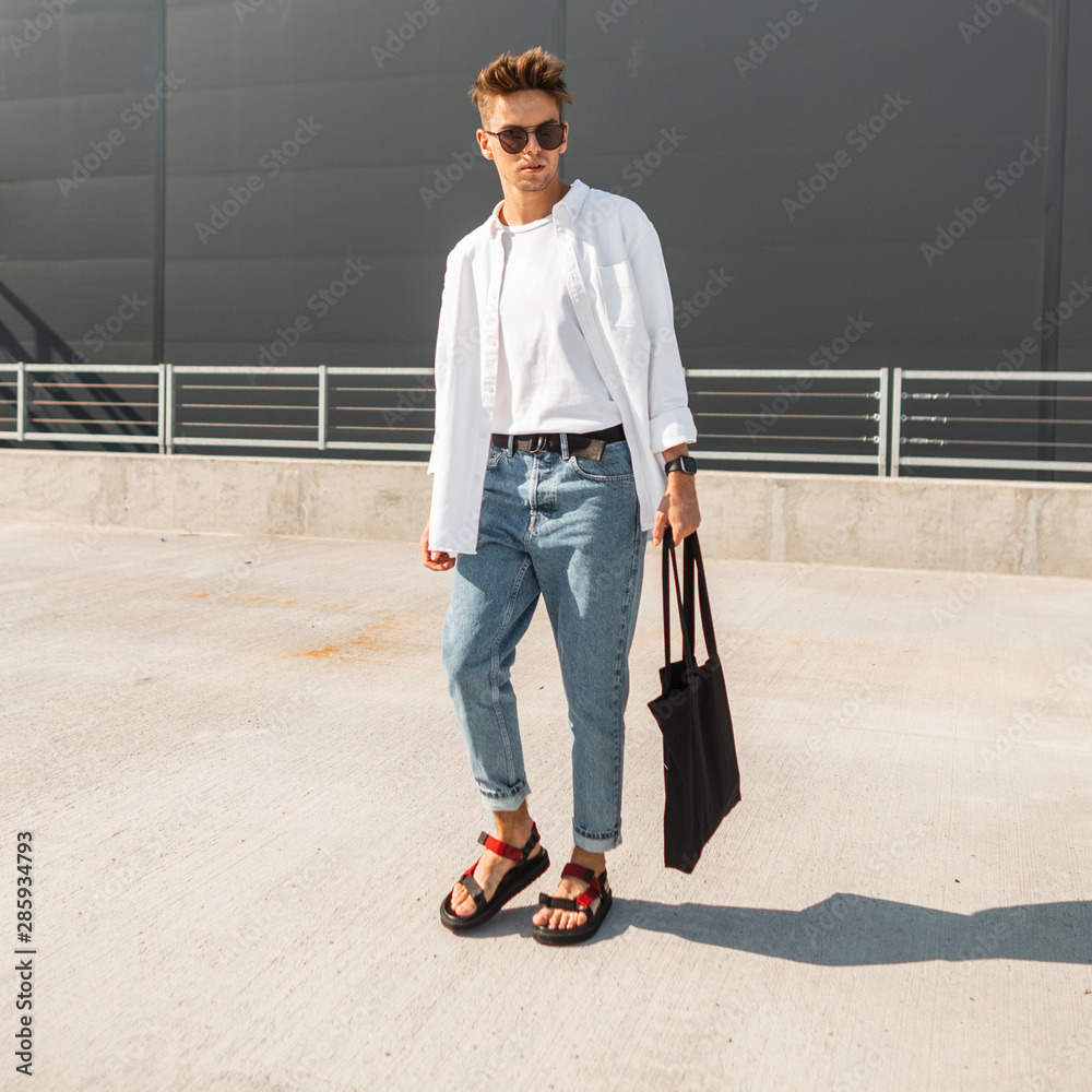 European young man hipster in a white shirt in blue fashionable jeans in  dark sunglasses with