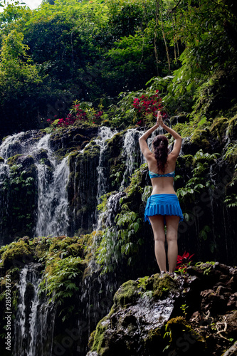 Young Caucasian woman meditating  practicing yoga at waterfall. View from back. Namaste mudra. Banyu Wana Amertha waterfall Wanagiri  Bali  Indonesia.