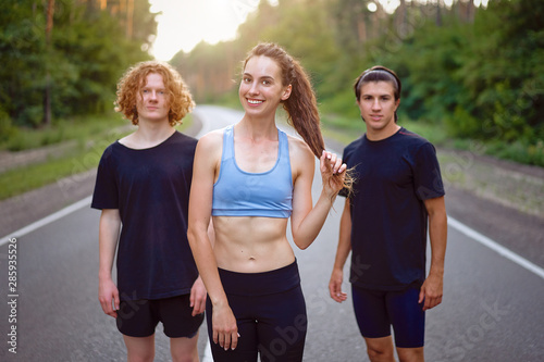 A group of three people athletes one girl and two men standing on asphalt road in pine forest at summer before jogging