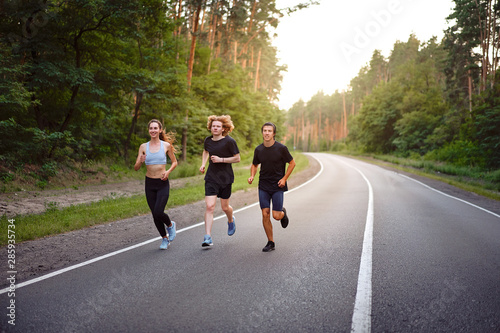 A group of three people athletes one girl and two men run on an asphalt road in a pine forest.