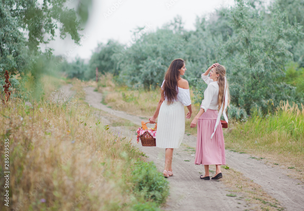 Two cheerful young women in retro style clothes are walking along the landing with a picnic basket and talking. Joint walk in nature. Back view Two cheerful young women in retro style clothes are walking along the landing with a picnic basket and talking. Joint walk in nature. Back view