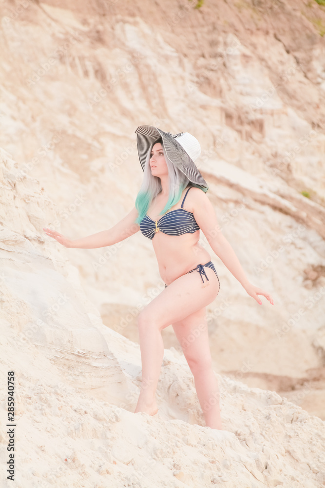 Young beautiful Caucasian woman in striped big hat with fashion swimwear posing in desert landscape with sand. Young beautiful Caucasian woman in striped big hat with fashion swimwear posing in desert landscape with sand.
