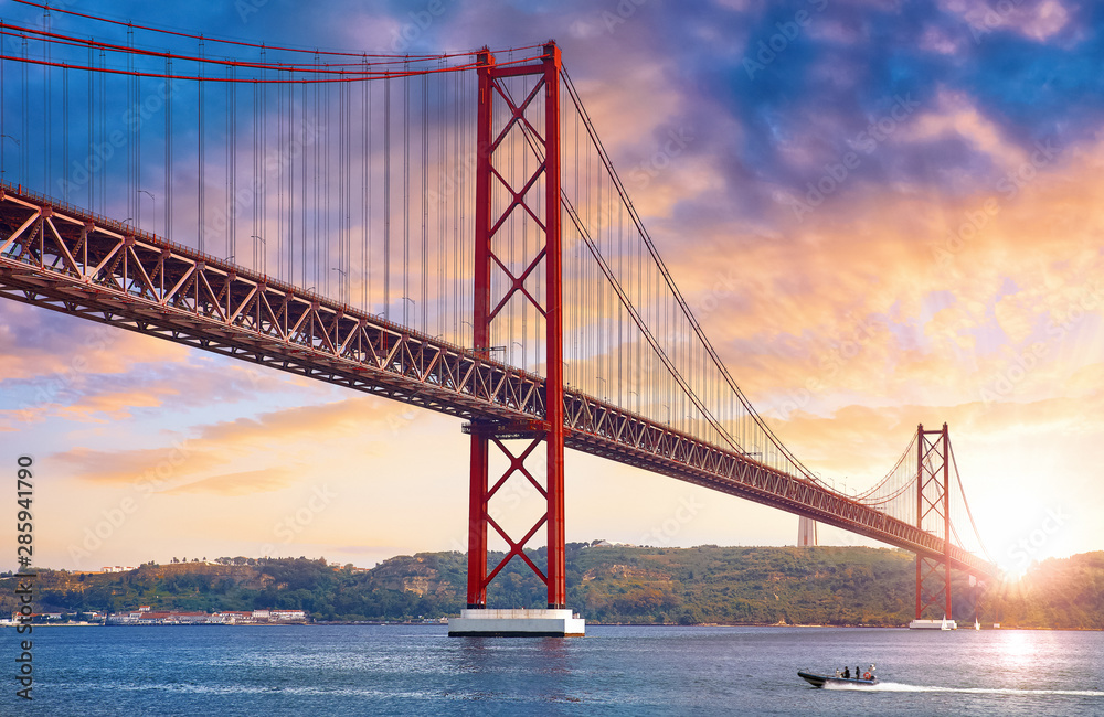 25th April Bridge in Lisbon, Portugal. Famous landmark on river Tagus. Summer sunny landscape with evening dusk sunset sky with clouds and sunlight. 25th April Bridge in Lisbon, Portugal. Famous landmark on river Tagus. Summer sunny landscape with evening dusk sunset sky with clouds and sunlight.