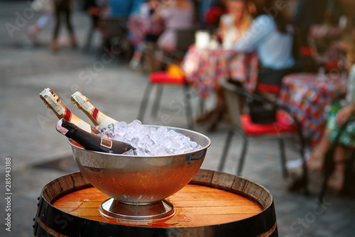 bottles of champagne in a bucket with ice stand on wooden barrel on a summer terrace. Restaurant guests in the background. Concept of social and cultural aspects of drinking. Restaurant business