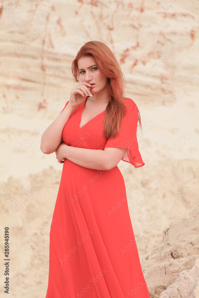 Fototapeta premium Young beautiful Caucasian woman in long red dress posing in desert landscape with sand.
