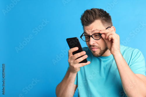 Man with vision problems using smartphone on blue background, space for text