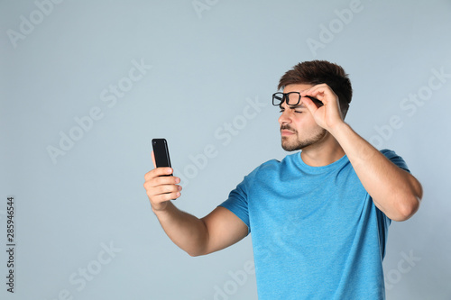 Young man with vision problems using smartphone on grey background, space for text