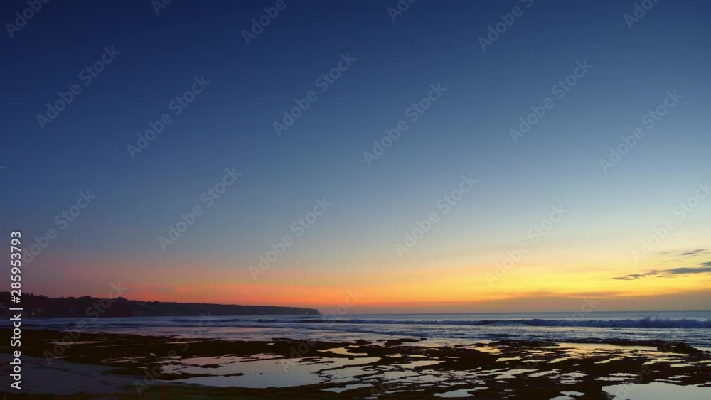 Panorama of a beach after sunset with dark blue and red sky during low tide with high waves on reef and rock Panorama of a beach after sunset with dark blue and red sky during low tide with high waves on reef and rock