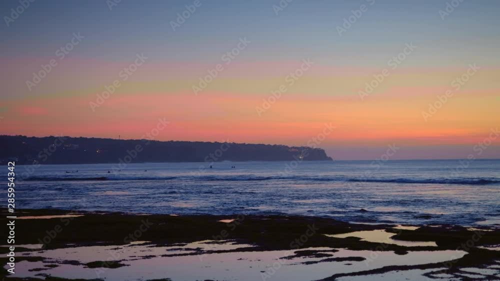 Panorama of a beach after sunset with surfers are swimming in dark blue water and waves near rock and cliff