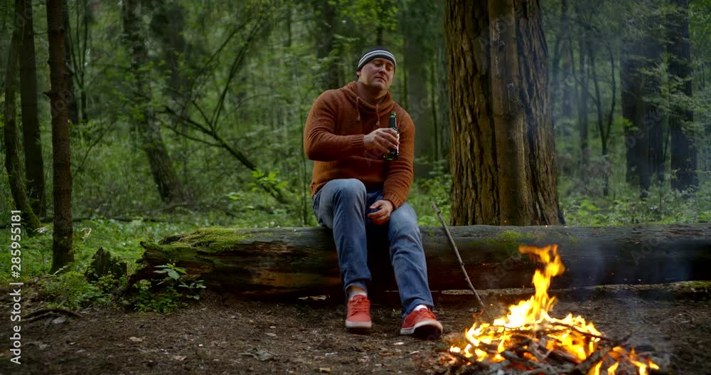 sorrowful and lonely man is drinking beer and looking on flame of campfire in woodland sorrowful and lonely man is drinking beer and looking on flame of campfire in woodland