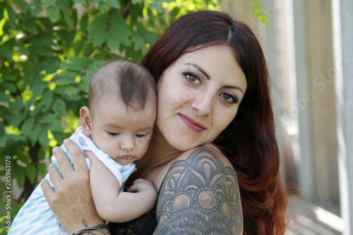 A young happy mother lovingly holds in her arms her 2-month-old son, a newborn, cute baby. Happy motherhood. Girl with a tattoo on her arm.
