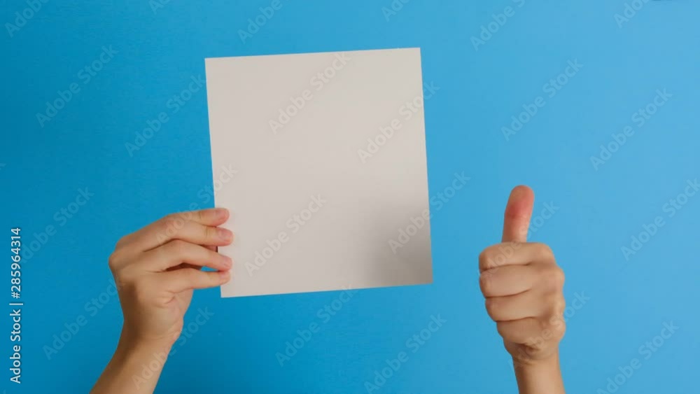 Female hands holding blank paper sheet isolated demonstrates approval or agreement on blue background with copy space. Body language concept. Hand sign. Like gesture Female hands holding blank paper sheet isolated demonstrates approval or agreement on blue background with copy space. Body language concept. Hand sign. Like gesture
