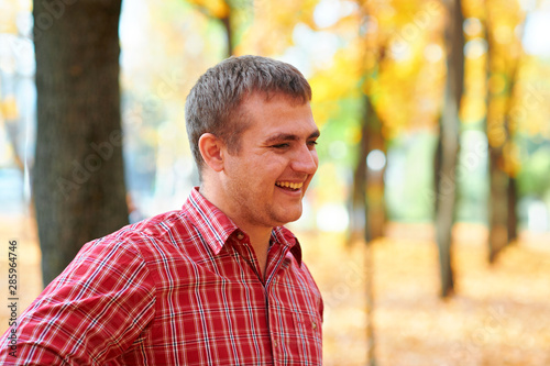 Portrait of happy man in autumn city park. Dressed in red plaid shirt. Bright yellow trees and leaves