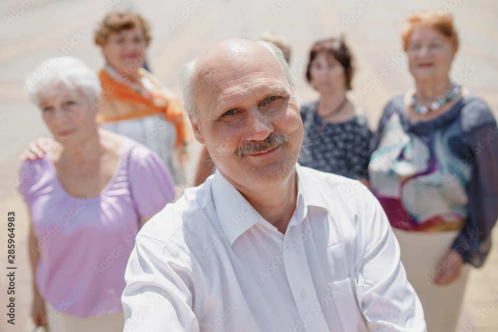 The group of smiling elderly people are making selfie together in a park on a warm day The group of smiling elderly people are making selfie together in a park on a warm day