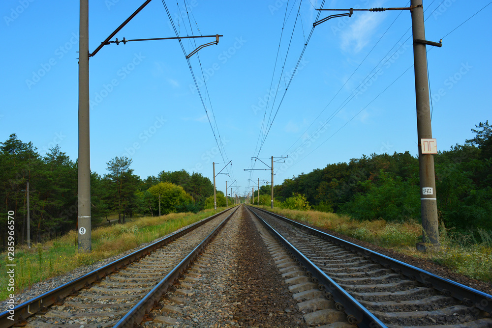 Railway track, railway and electric power lines for electric vehicles, electric trains lying along coniferous forests with blue sky, clouds. Infrastructure of Severny housing estate, Dnipro, Ukraine.