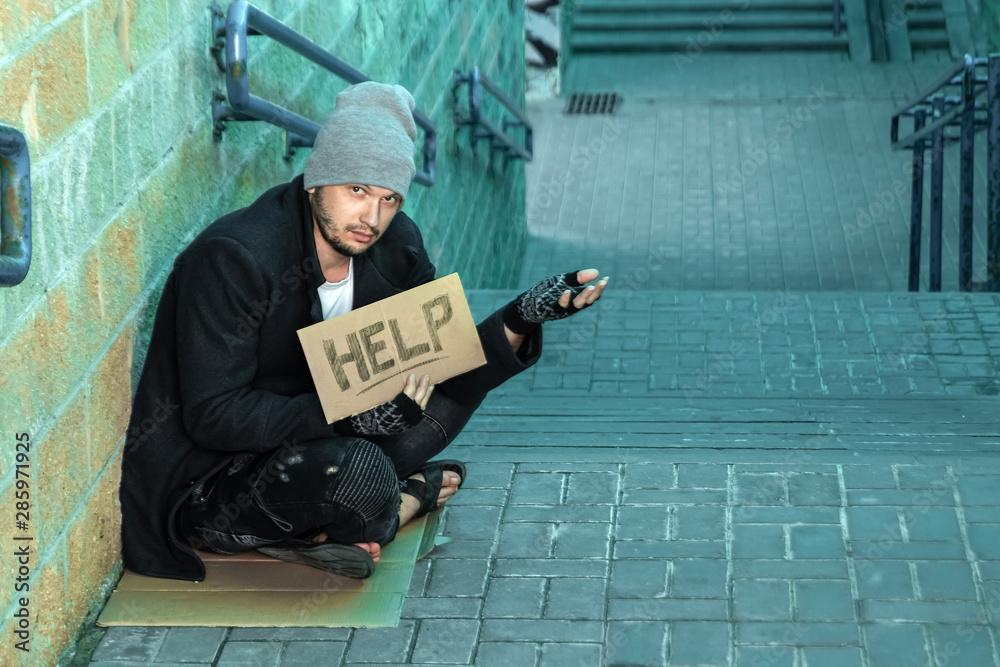 A man, homeless, a person asks for alms on the street with a Help sign. Concept of homeless person, addict, poverty, despair. A man, homeless, a person asks for alms on the street with a Help sign. Concept of homeless person, addict, poverty, despair.