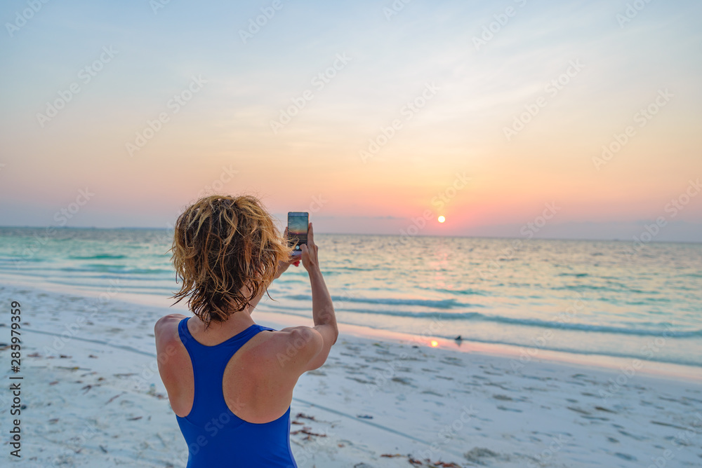 Woman taking photo with smartphone of romantic sky at sunset on sand beach, rear view, real people traveling around the world. Indonesia tropical destination, lifestyle sharing social media concept.