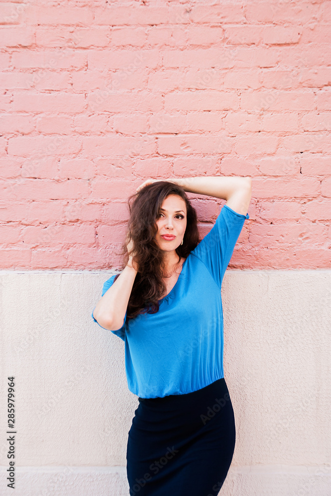 A beautiful brunette woman in a blue blouse posing against a textured pink brick wall. Portrait of a young woman A beautiful brunette woman in a blue blouse posing against a textured pink brick wall. Portrait of a young woman