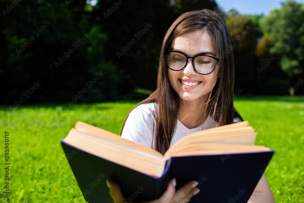 Fototapeta premium Pretty brunette girl in glasses reading book in park close-up