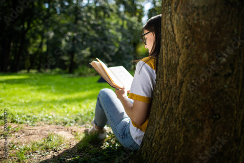 Smiling young woman in jeans reading book resting against park tree view from behind