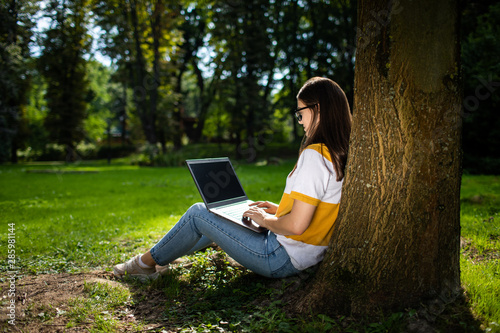 Young woman working by laptop in park leaning against a tree