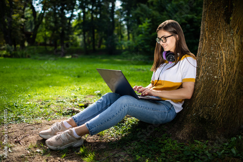 Smiling cute girl using a laptop resting against a park tree