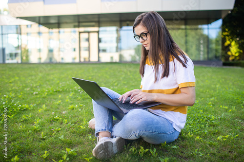 Pretty thoughtful student girl studying by laptop on a lawn in front of the college © proimagecontent