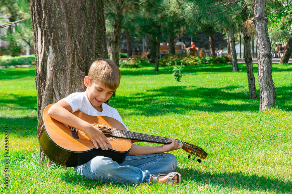 Fototapeta premium A boy with a guitar sits under a tree, sings songs and enjoys nature.