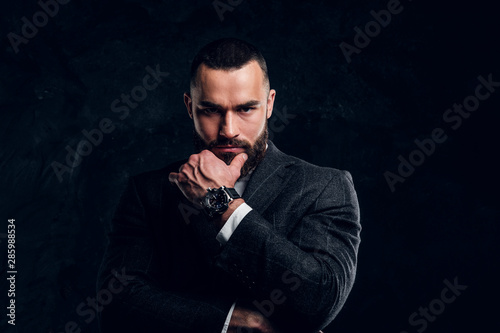Serious sceptical buisnessman with beard and in dark clothing is standing at dark photo studio.