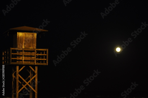 Caseta de guardavidas hecha de madera en la costa de la una playa española en horario nocturno, la luz de la luna deslumbrante en un costado, cielo azul oscuro, azul marino