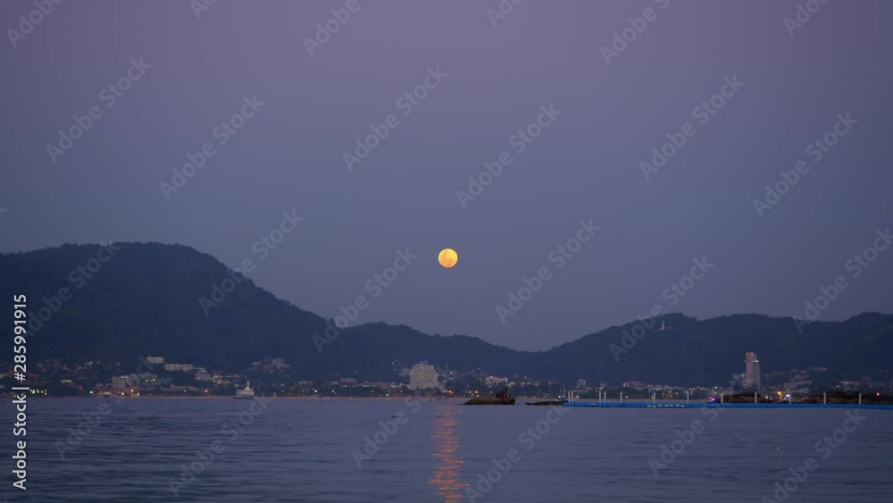 Panorama of a city from beach with small waves and calm sea with hills and big full moon in background Panorama of a city from beach with small waves and calm sea with hills and big full moon in background