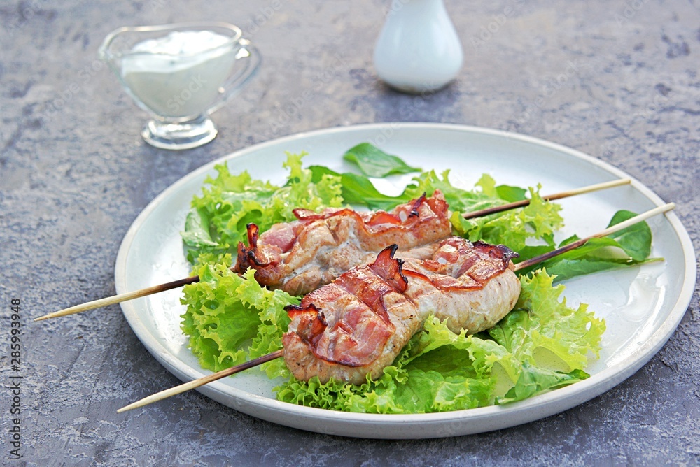Grilled pork with bacon on wooden skewers, on a white ceramic tray on a gray concrete background. Served with lettuce. Barbecue. Selective focus. Grilled pork with bacon on wooden skewers, on a white ceramic tray on a gray concrete background. Served with lettuce. Barbecue. Selective focus.