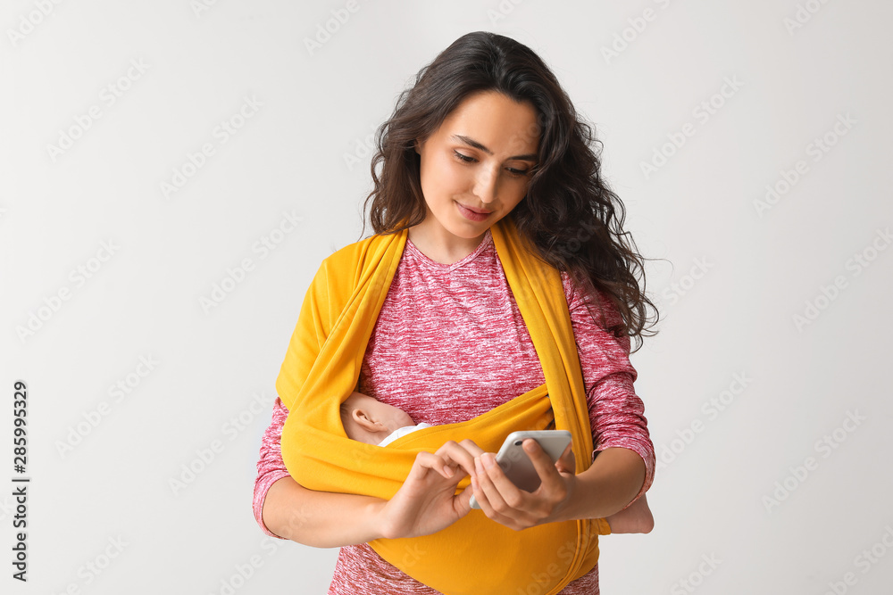 Young mother with little baby in sling using mobile phone on light background Young mother with little baby in sling using mobile phone on light background