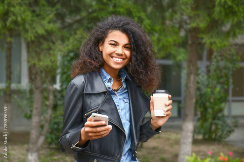 Beautiful young African-American woman with coffee and mobile phone outdoors