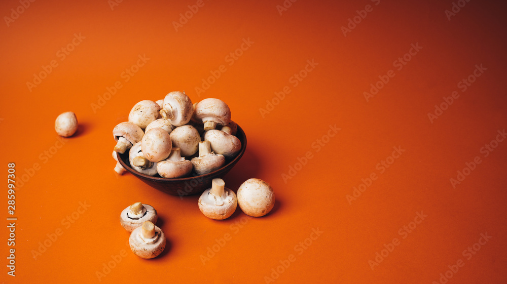 Mushrooms in a wooden bowl on an orange background. The small white champignon in a plate and scattered near it.