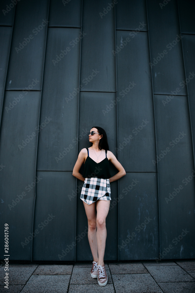 Fototapeta premium Sexy girl near a dark wall in a T-shirt, sunglasses and short shorts