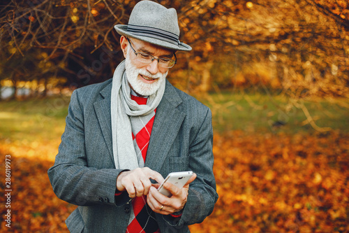 Handsome grandfather in a autumn park. Old man in a gray jacket and hat. Male with mobile phone