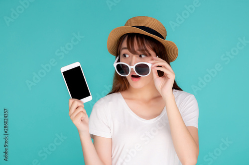 Beautiful young asian woman presenting and showing smart mobile phone with surprise in vacation isolated blue background, girl excited on smartphone in travel summer trip, communication concept.