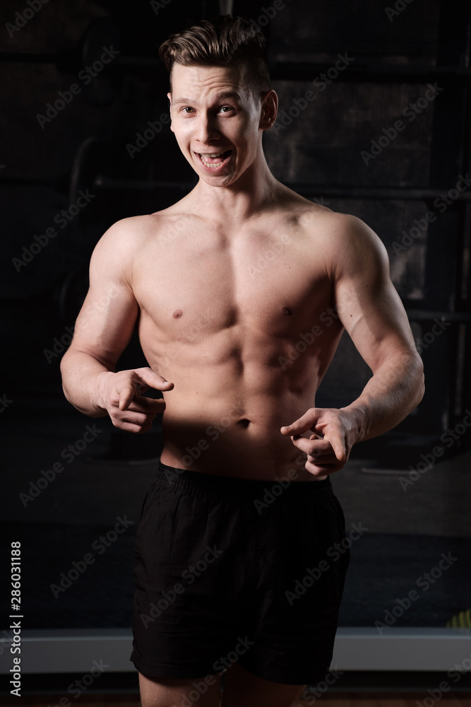 A muscular guy with a naked torso is standing in the gym and smiling, looking into the frame while holding index fingers forward. A muscular guy with a naked torso is standing in the gym and smiling, looking into the frame while holding index fingers forward.