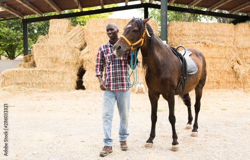 Young man farmer standing with horse at stable at farm