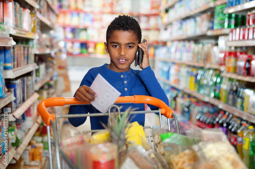 Focused African tween boy talking on phone and looking at shopping list while visiting supermarket