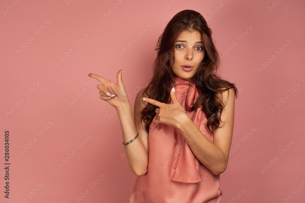 A dark-haired girl in a pink dress is standing on a pink background, pointing with her index fingers to the side, raising eyebrows A dark-haired girl in a pink dress is standing on a pink background, pointing with her index fingers to the side, raising eyebrows