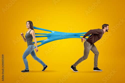 Young brunette girl wearing casual jeans and t-shirt stuck to young man in casual clothes with blue sticky slime on yellow background.